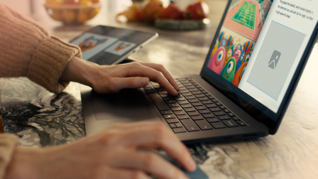 An image of a hand on a mouse and fingers the keyboard of a Surface Laptop with a Surface Pro in the background