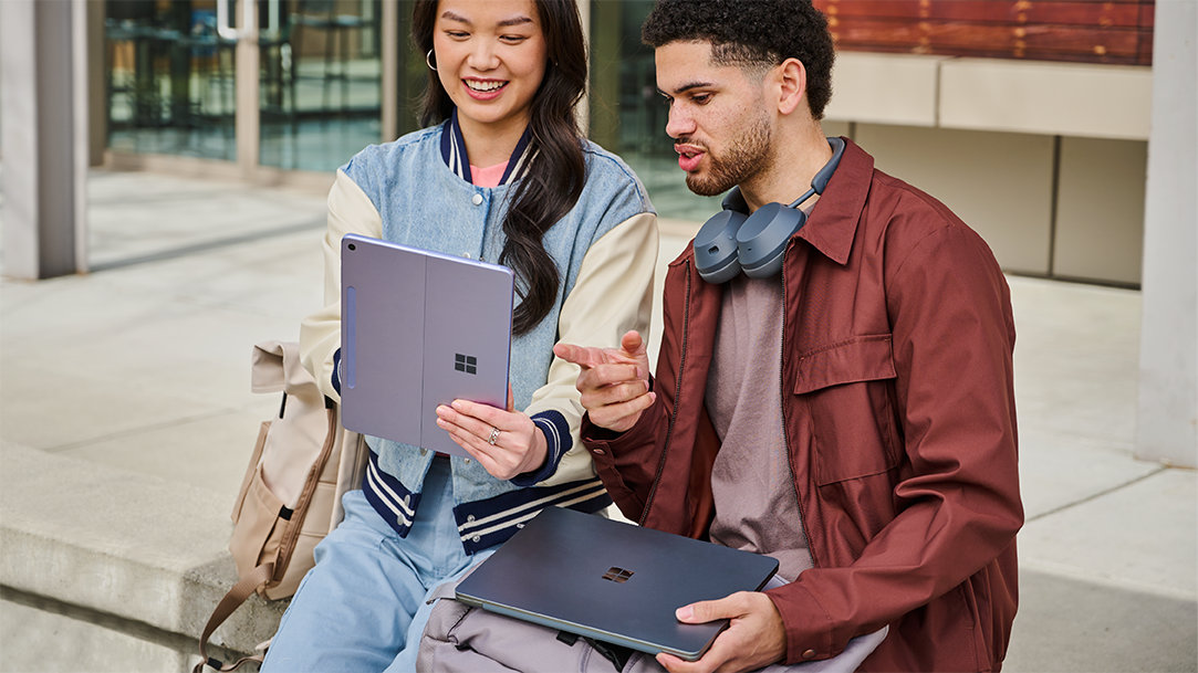 An image of a woman holding a Surface Pro and a man holding a Surface Laptop, sitting outside of a building