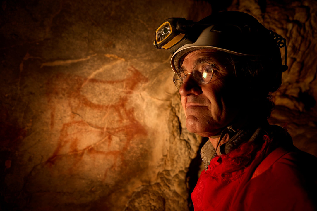 Archeologist stands inside a remote Spanish cave in front of a cave drawing in red ochre of a deer or ibex