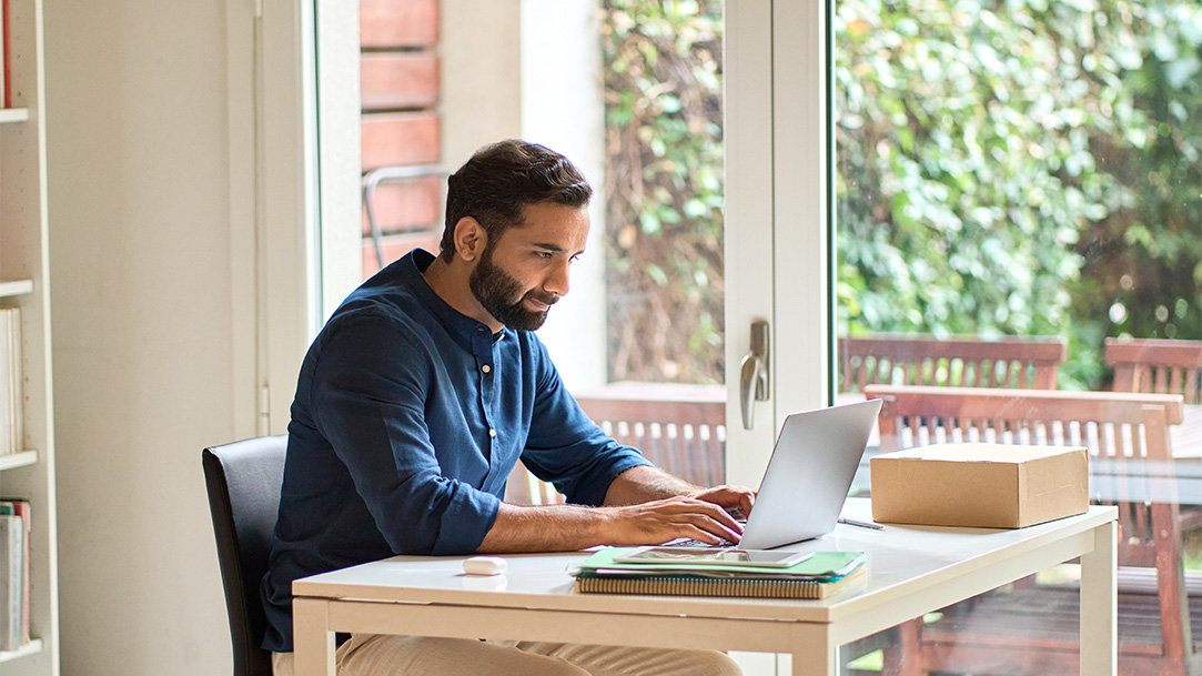 Remote worker sitting at a home desk with an open Windows 11 Pro device, notebooks, and a view onto a deck with table and chairs