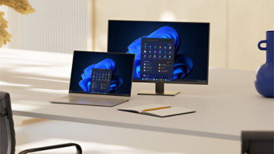 An open laptop and monitor sit on a desk alongside a notebook and pencil, with both screens displaying Windows blue bloom backgrounds and open start menus