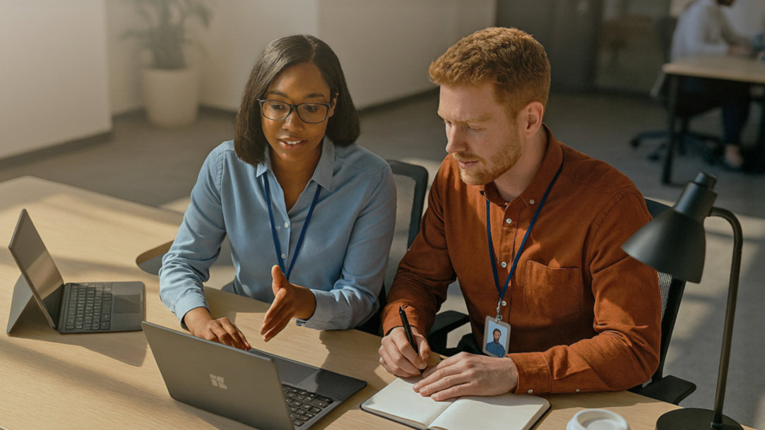 Two office workers sitting together at a desk, each with an open Windows Copilot+ PC laptop, notebooks, and a lamp