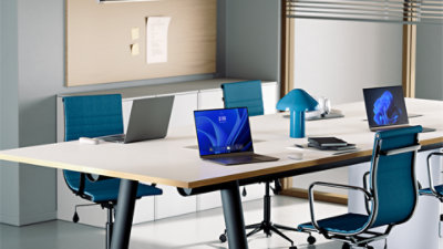 Three open laptops sit on a conference table surrounded by blue office chairs, with one laptop displaying a Windows welcome screen and one laptop displaying a Windows bloom background