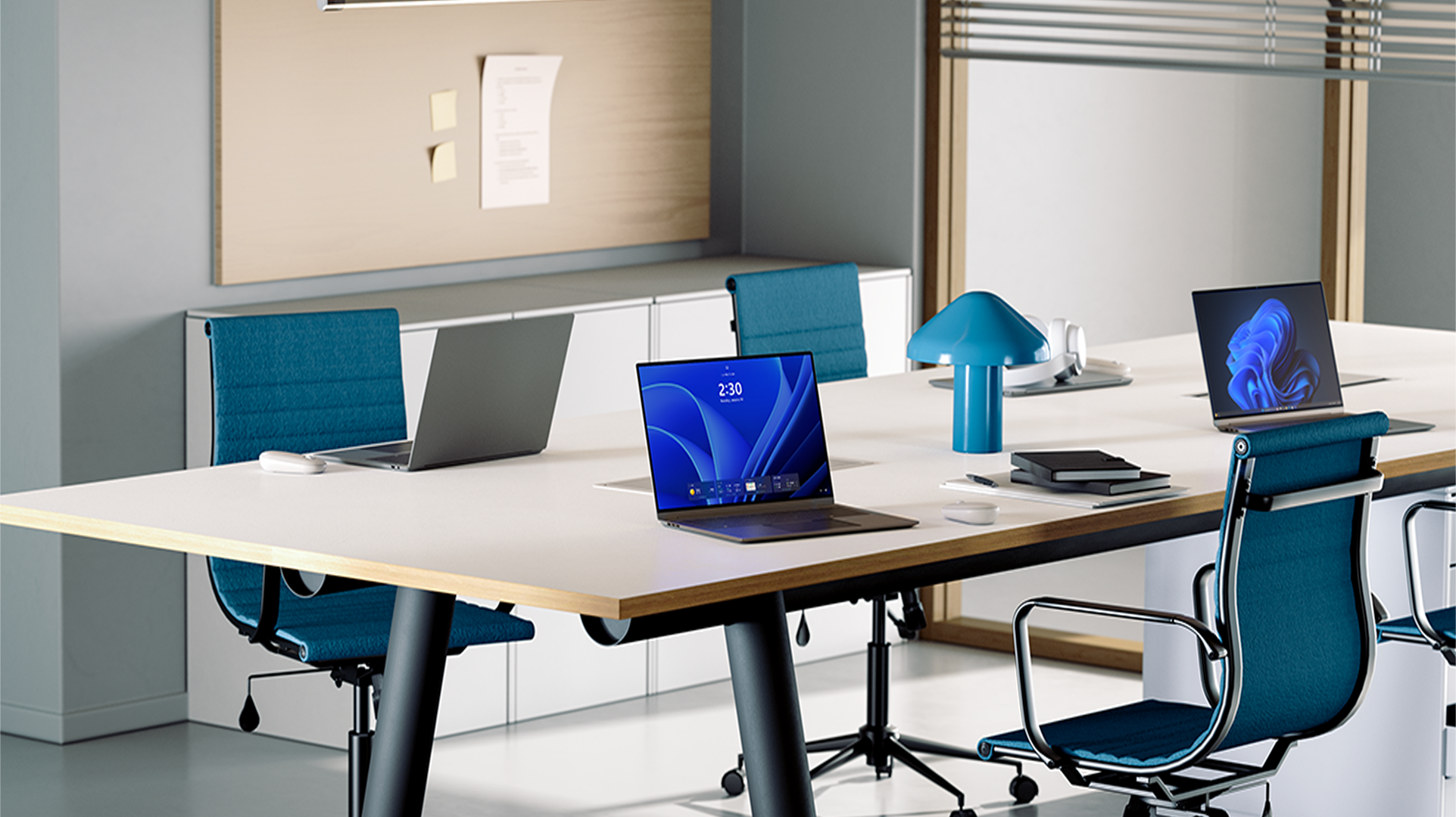 Three open laptops sit on a conference table surrounded by blue office chairs, with one laptop displaying a Windows welcome screen and one laptop displaying a Windows bloom background