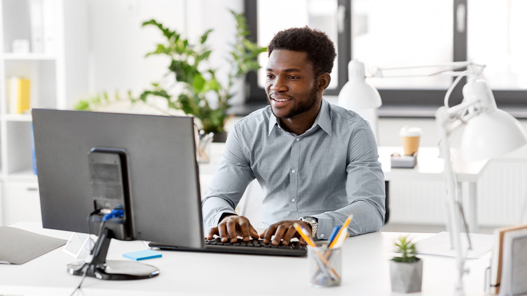 An office worker sitting at a workstation desk with a Windows 11 Pro PC device, a keybaord, pen cup, desk lamp, and another empty workstation in the background