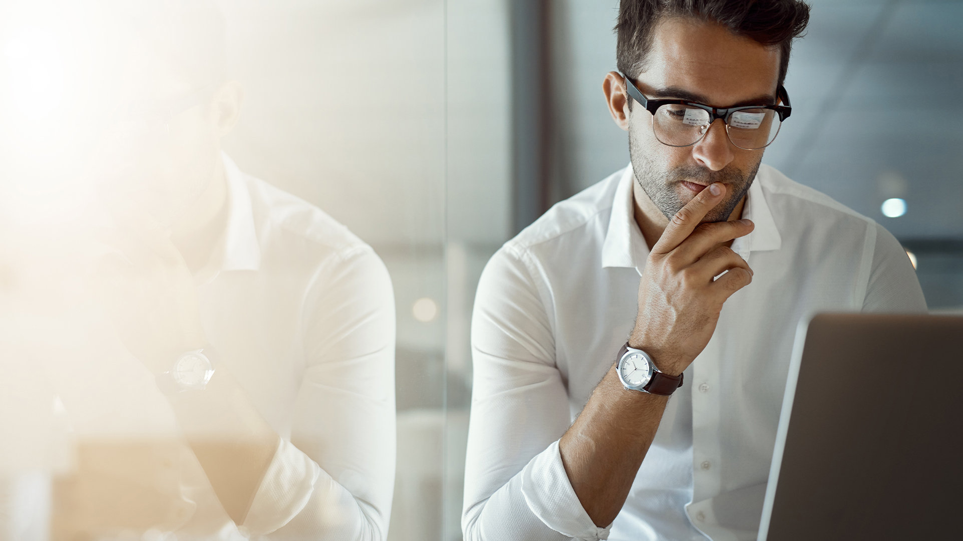 Office worker sitting at a desk in a brightly lit office with a large window, reading from an open laptop on a desk