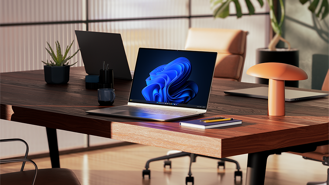 Three Windows 11 Pro PCs, one open and displaying the bloom image, all sitting on a wood desk, with a notebook, pen, a plant, a lamp, and office chairs