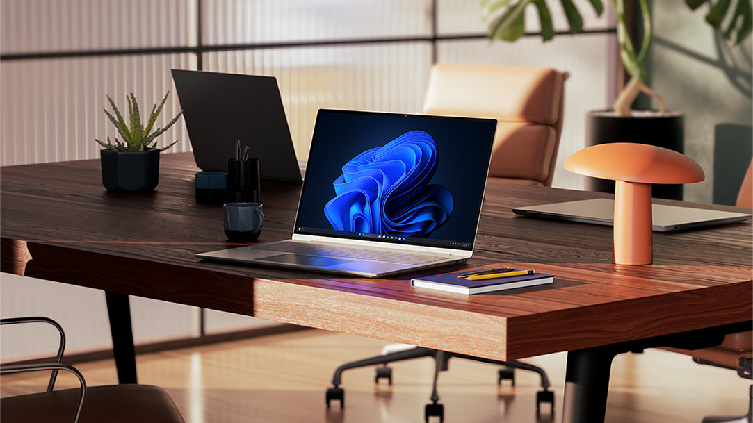 Three Windows 11 Pro PCs with one open and displaying the bloom image, all sitting on a wood desk, with a notebook, pen, a plant, and office chairs