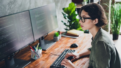 Woman in a green shirt wearing glasses and sitting at a desk typing on a keyboard while looking at two monitors full of code