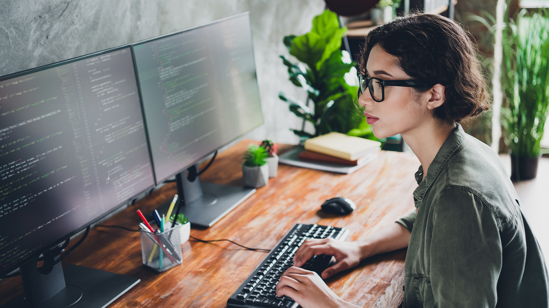 A seated office worker with a brightly lit workstation, a wood desk with two large monitors, a keyboard, mouse, a cup of pens, and several office plants