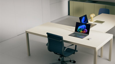 An open laptop displaying a Windows bloom background sits alongside a mouse and notebook on a white desk with a blue chair