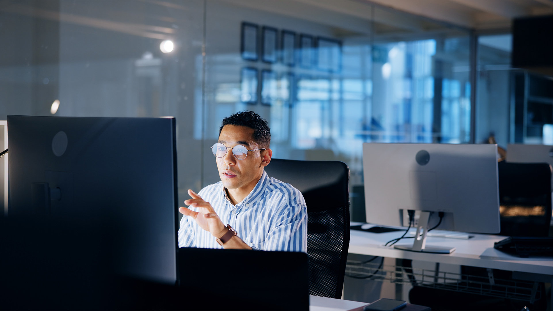 Office worker sitting at a workstation working at a large monitor, with a glass wall behind the desk, and other empty workstations in the background in a large open office area in late afternoon light