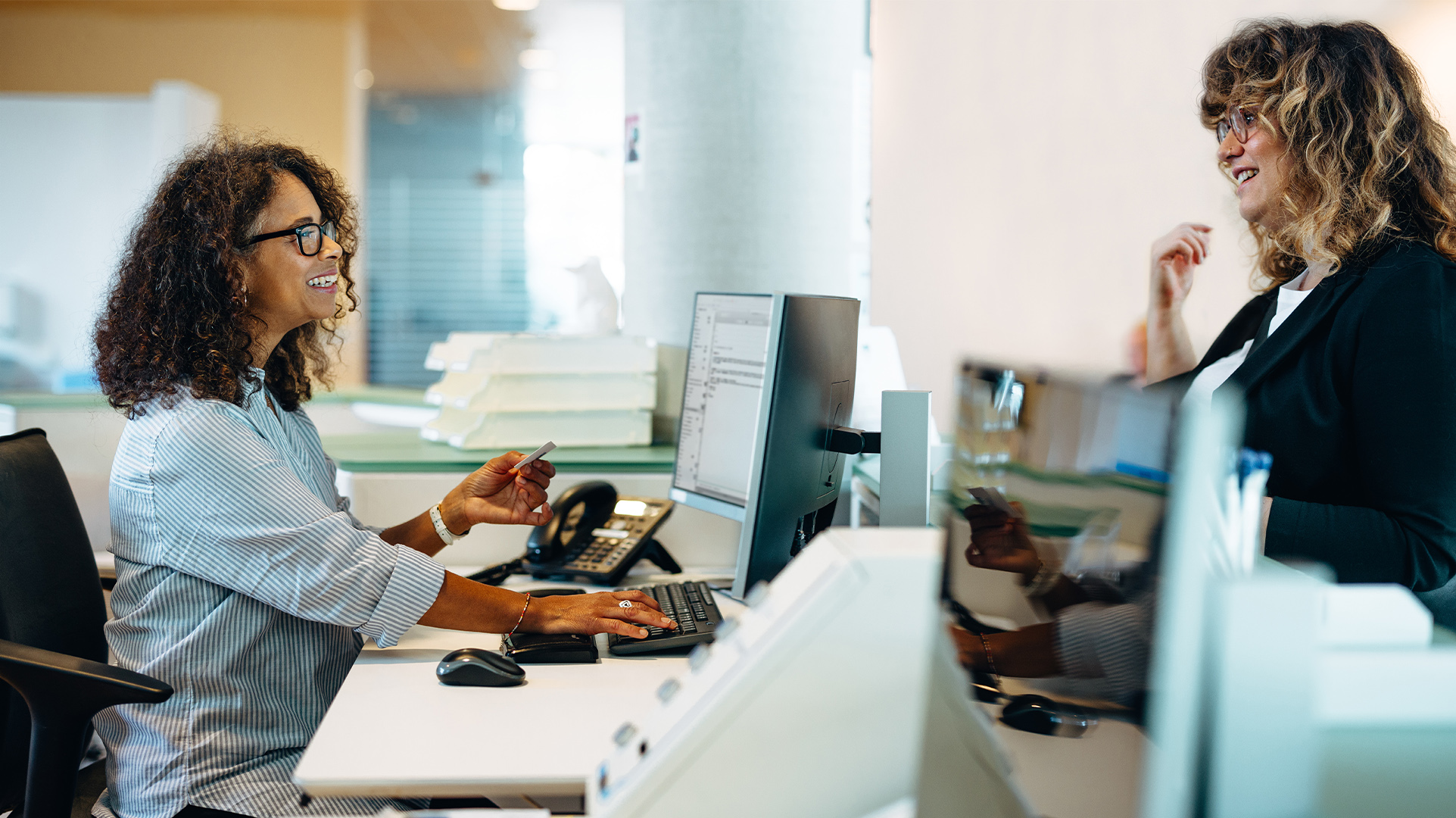 Two office workers at desks in a modern, open office setting with multiple workstations talking to each other, with phones, large monitors, and other office tools and supplies