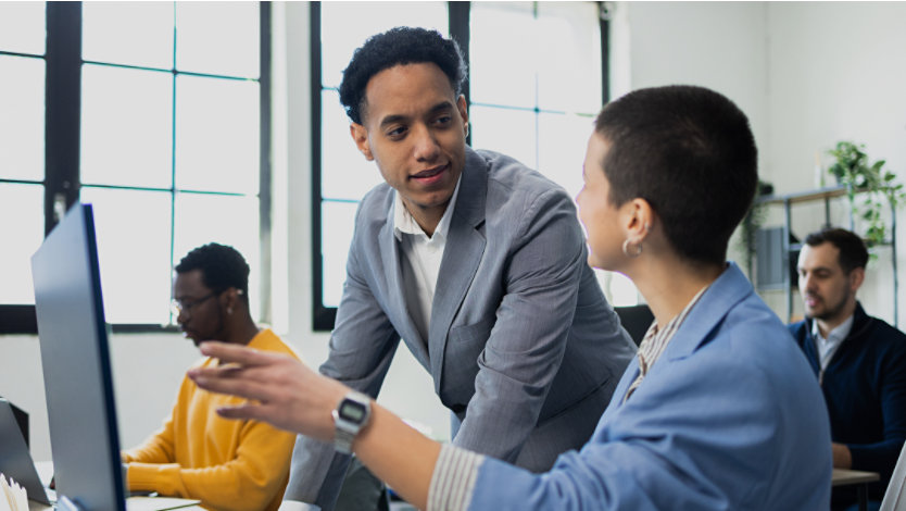 Four office workers in an open office setting, with two colleagues discussing something over an open laptop computer screen