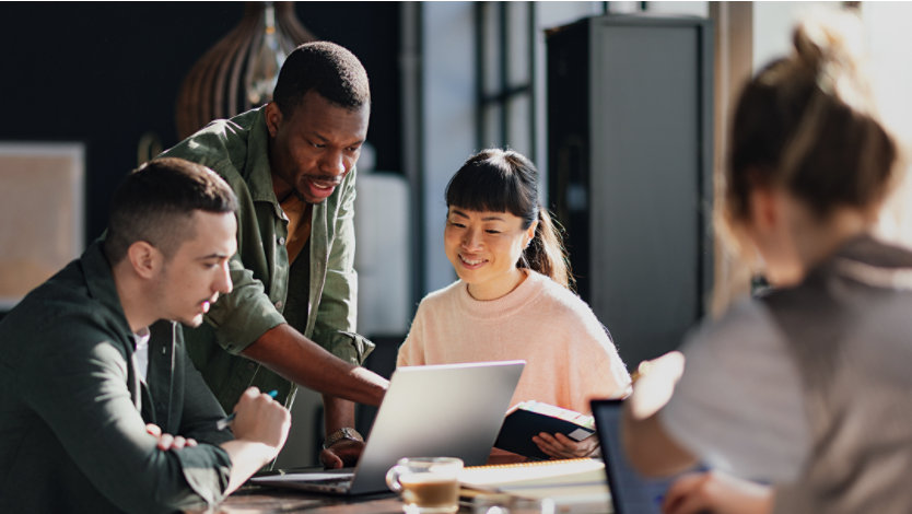 Four office workers sitting around a meeting table, with three discussing something on an open laptop screen