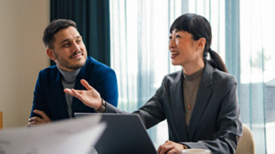 Ein Mann und eine Frau mit Anzug und Kostüm sitzen an einem Konferenztisch und arbeiten an einem Computer.