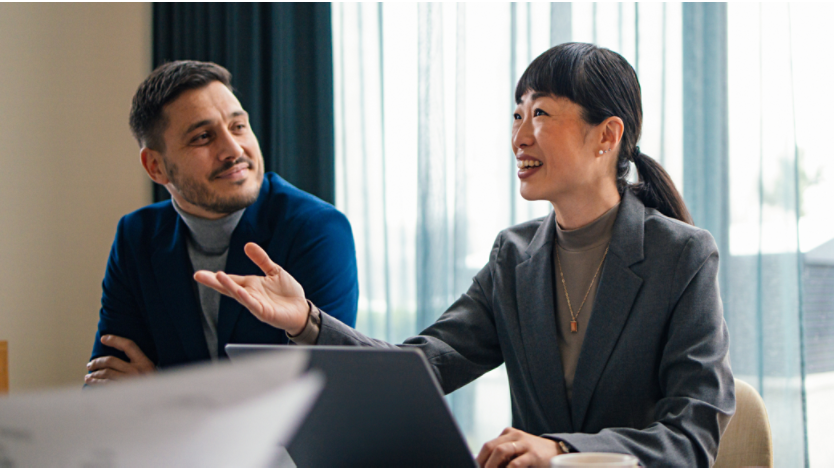 A man and woman wearing suites and sitting at a conference table while working on a computer.