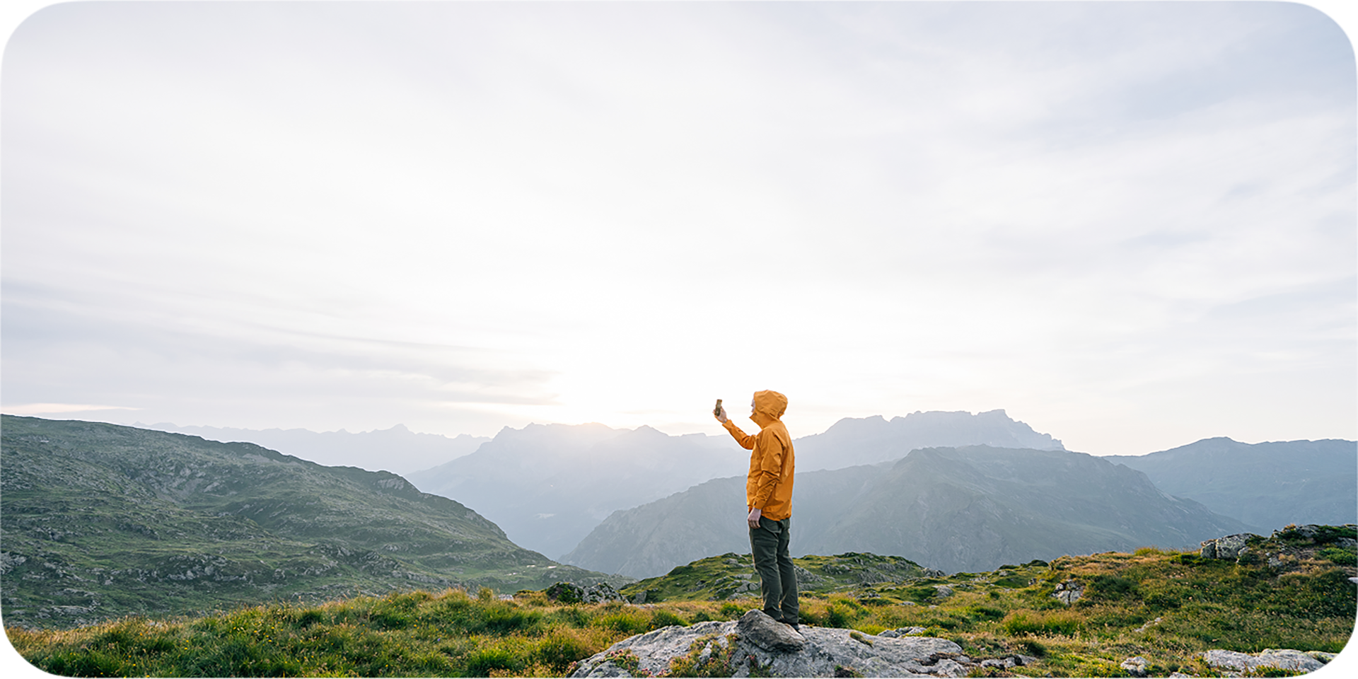 Person in a yellow jacket standing on a rocky hilltop, holding a phone against a backdrop of green valleys and distant mountain peaks under a bright sky
