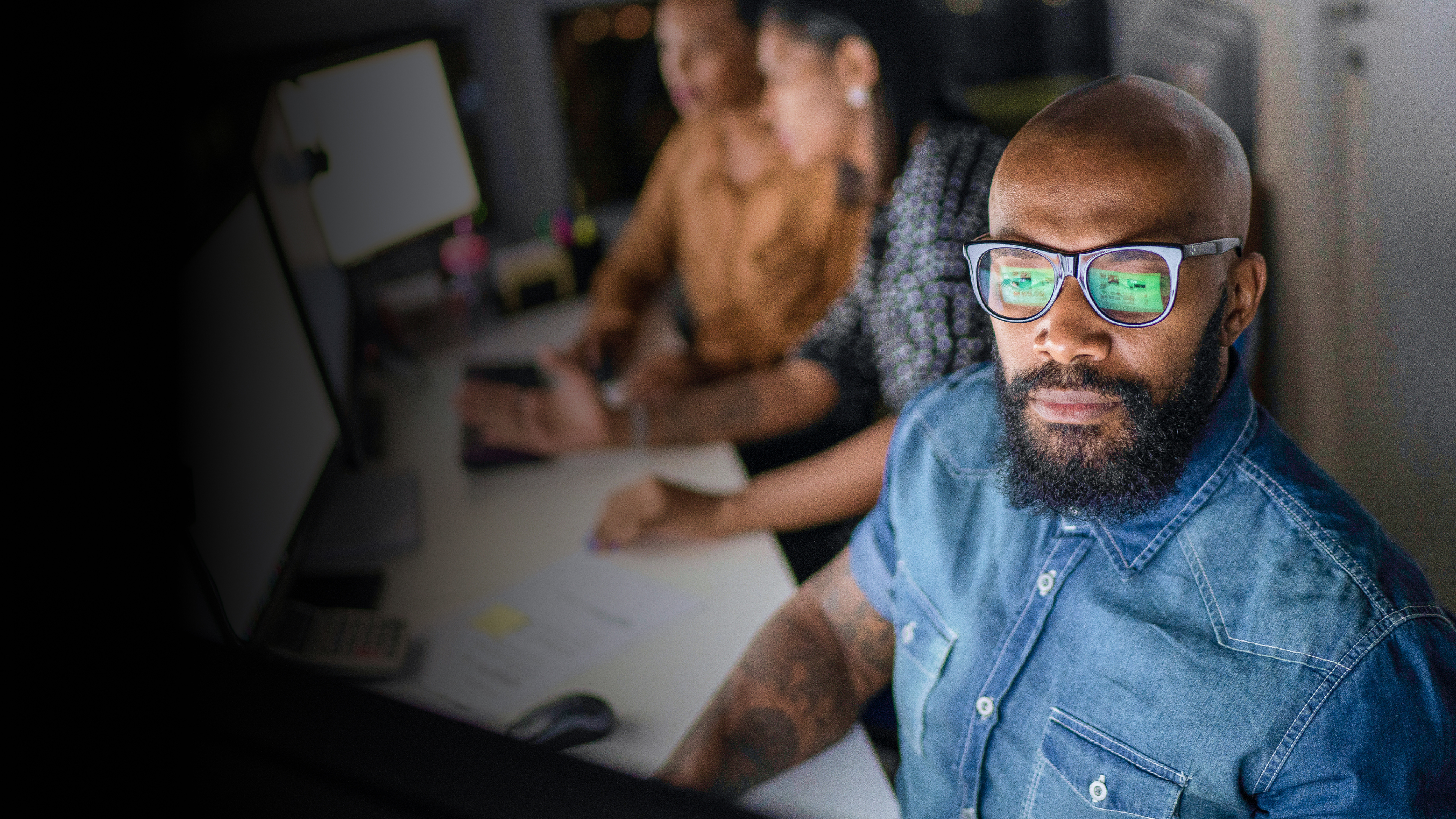 A man in a denim shirt is looking at his computer screen while two women work on the same computer behind him.