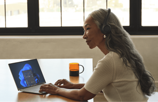 A woman with long gray hair wearing headphones, using a laptop at a table with an orange mug, in a sunlit room with large windows