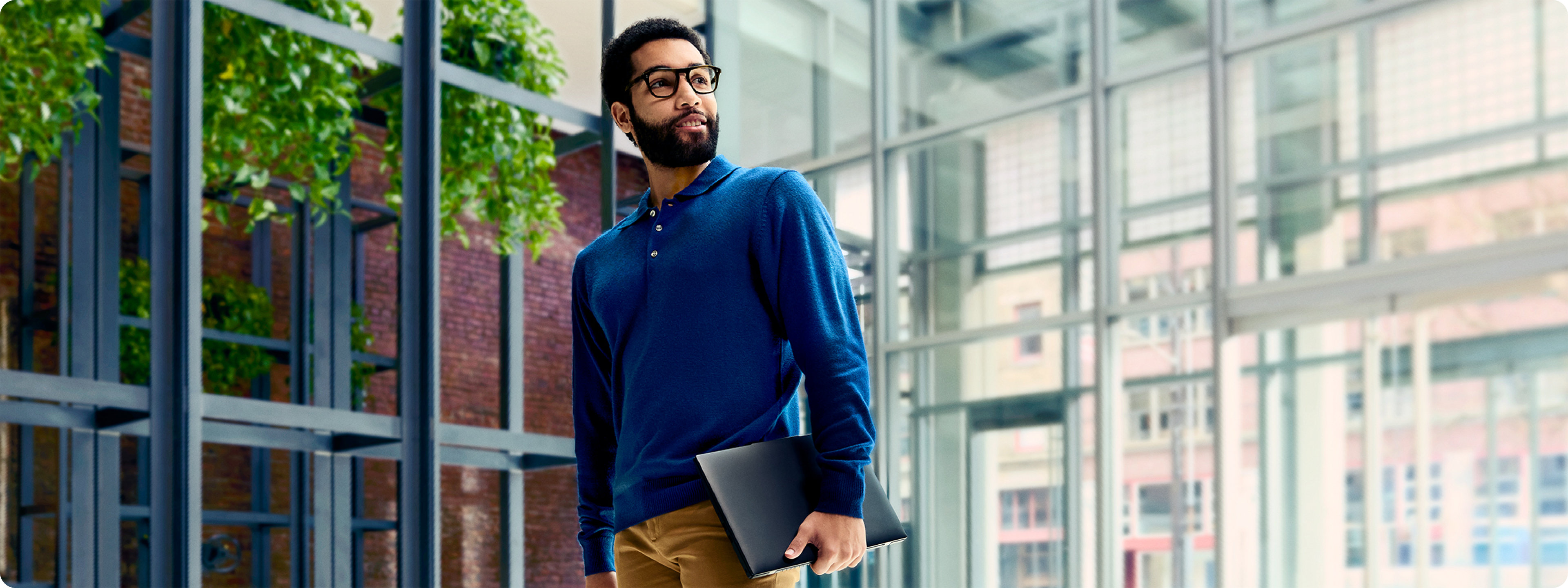 A man wearing a blue sweater is working on his laptop device while walking through a building with glass doors and windows.