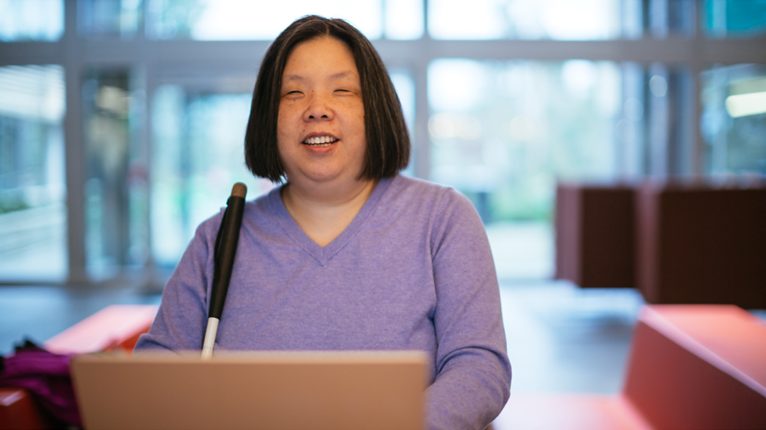 A woman in a purple top holding a white cane, seated at a laptop in a room with large windows and red furniture. 