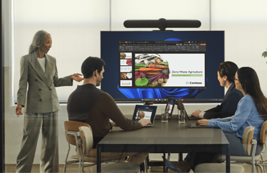 Four people in a meeting room, one presenting a slide while others sit with laptops.
