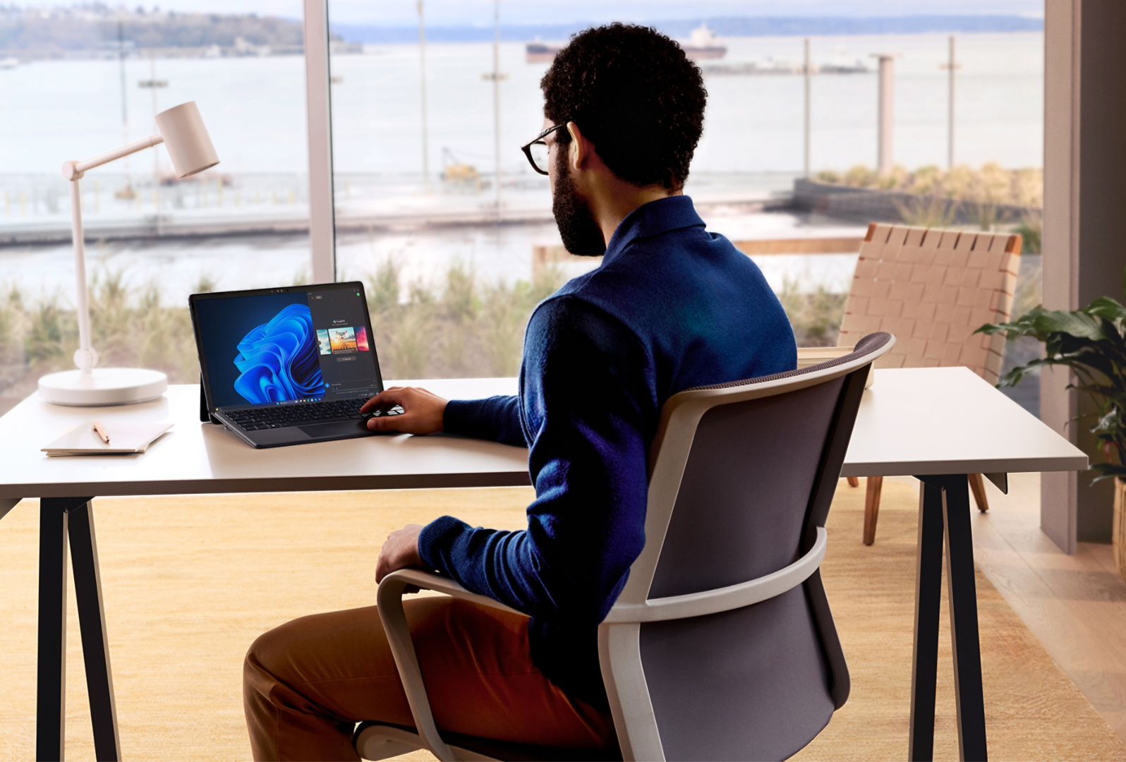 A man is sitting and working on a Microsoft 2-in-1 device from an office setting looking out at the water.