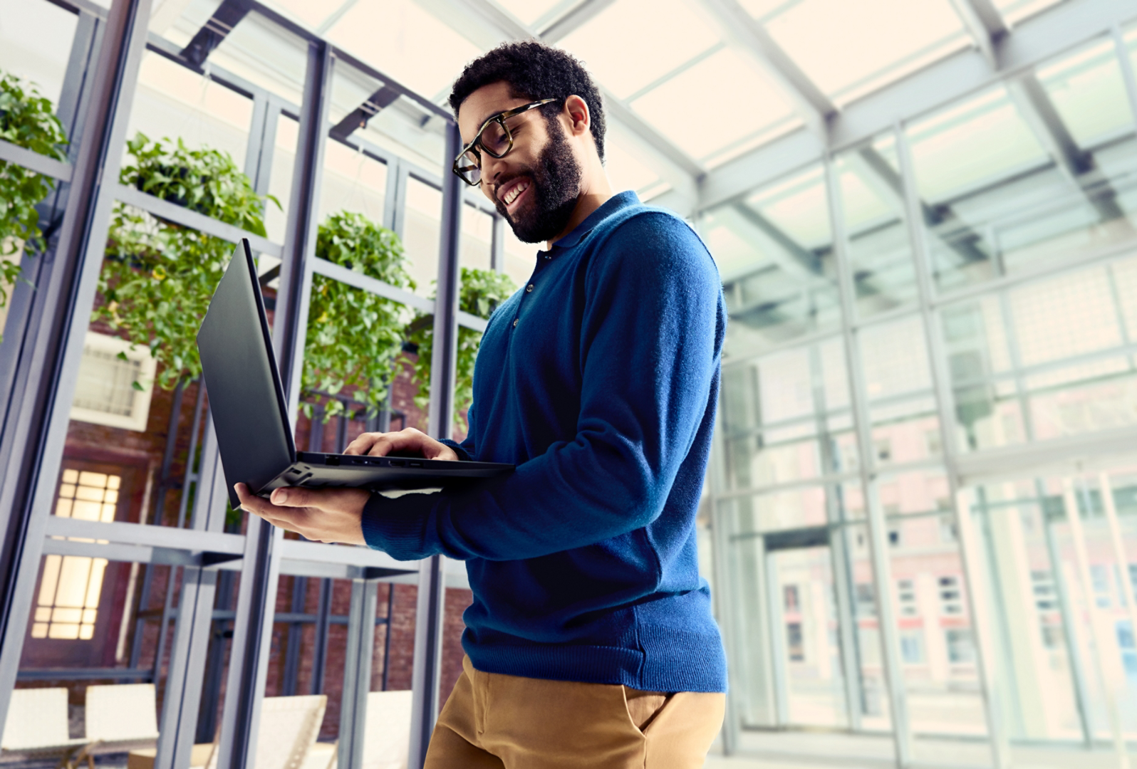 A man wearing a blue sweater is working on his laptop device while walking through a building with glass doors and windows.