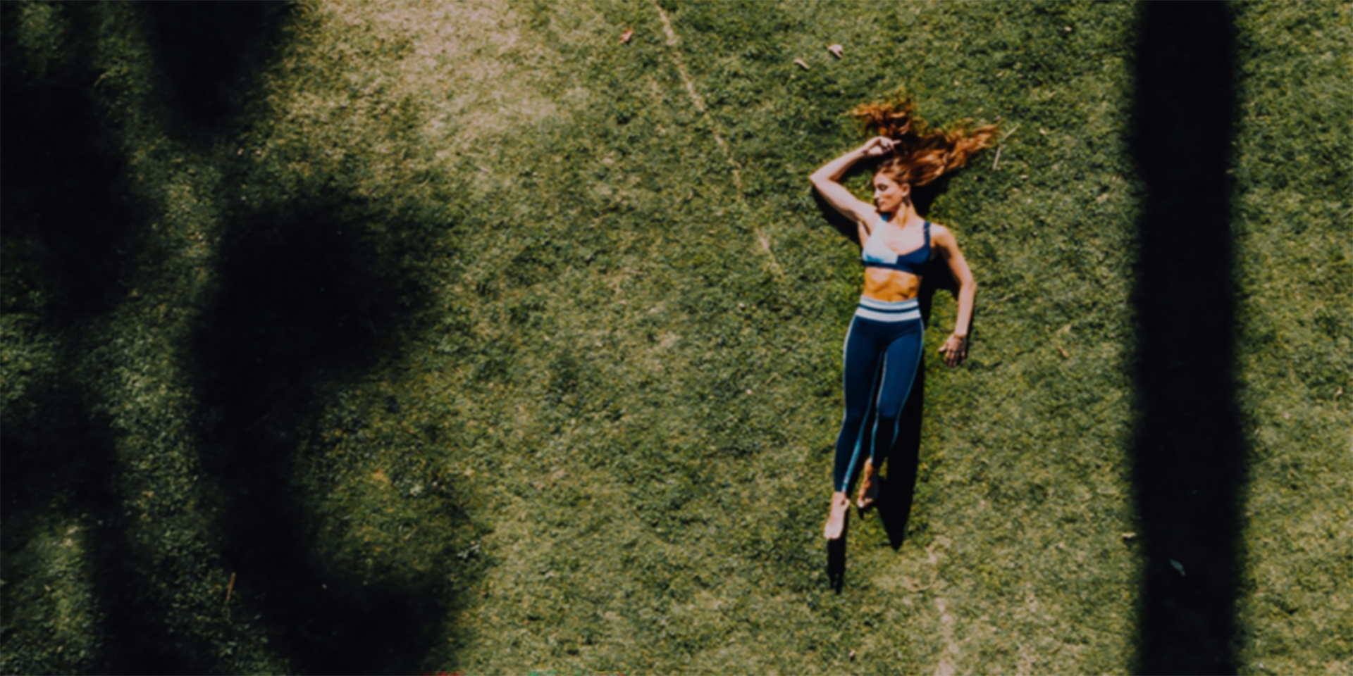 Person lying on green grass in athletic wear, viewed from above, with long shadows stretching across the scene under bright sunlight