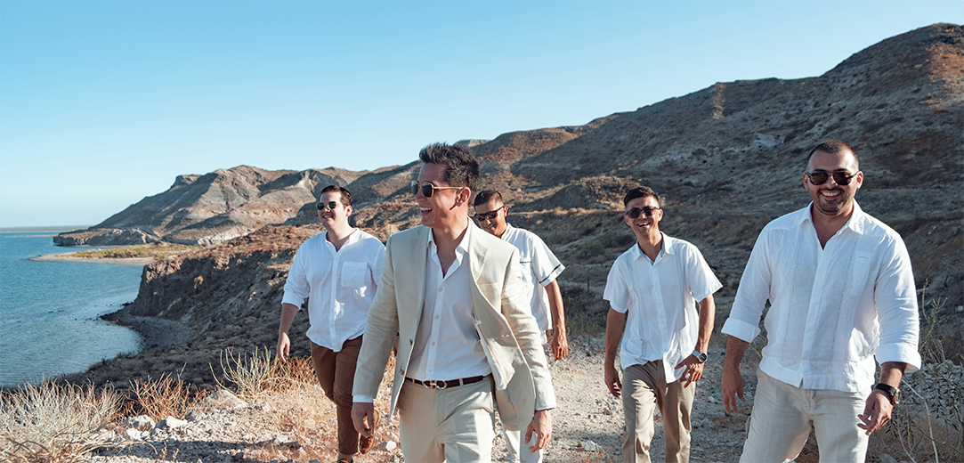 Five men smile while walking on a rocky trail above the ocean during the day