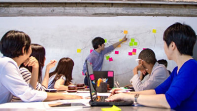 Five people sitting at a conference room table watching a sixth person who is standing and pointing to a drawing on a white board while one of the sitting people takes notes on a laptop