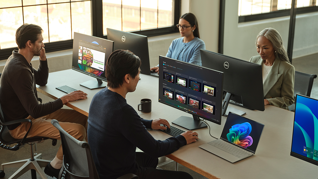 Four people sitting at a desk next to a window in an office working on Dell computers with external monitors