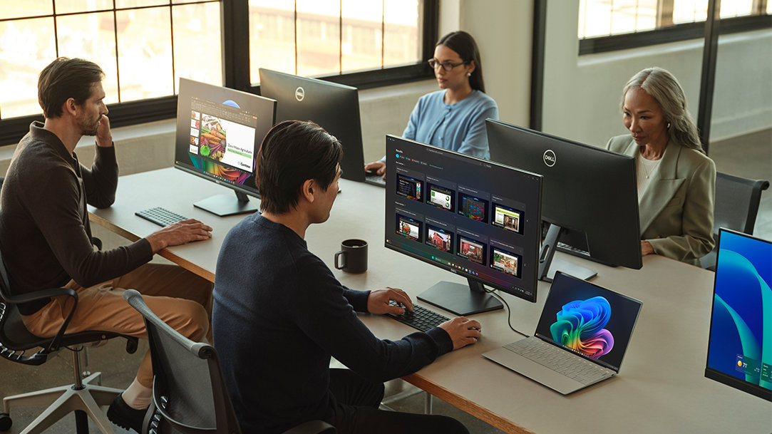 Four people sitting at a desk next to a window in an office working on Dell computers with external monitors