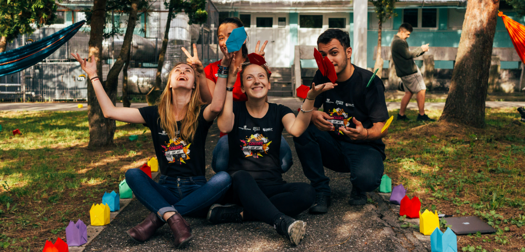 Four students sitting on the ground while smiling and throwing colorful paper in the air