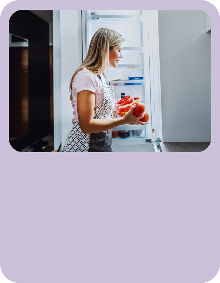 Person wearing a light pink shirt and patterned apron holding a bowl of tomatoes while standing in front of an open refrigerator