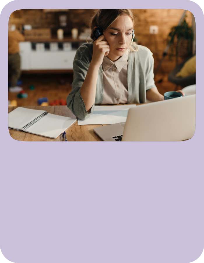 Person sitting at a wooden desk with an open laptop, notebook, and papers, holding a phone to the ear and a mug in the other hand