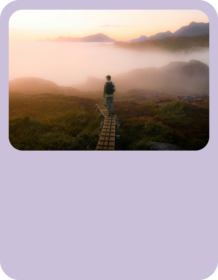 Person standing on a narrow wooden path overlooking a misty valley at sunrise, with mountains faintly visible in the background