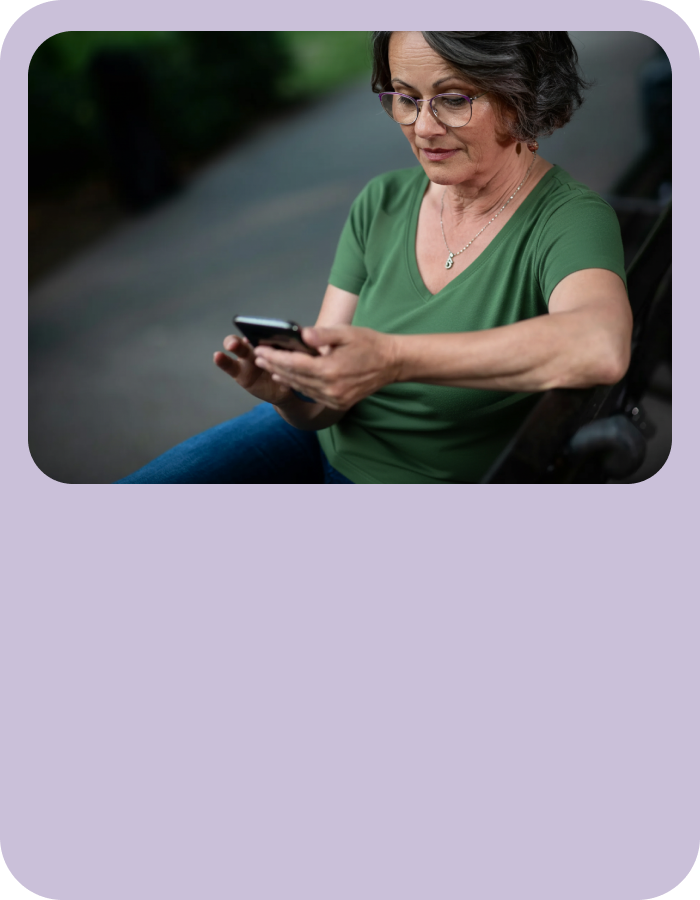 A person sitting outdoors on a bench, looking down at a mobile device, with soft natural light and greenery in the background.