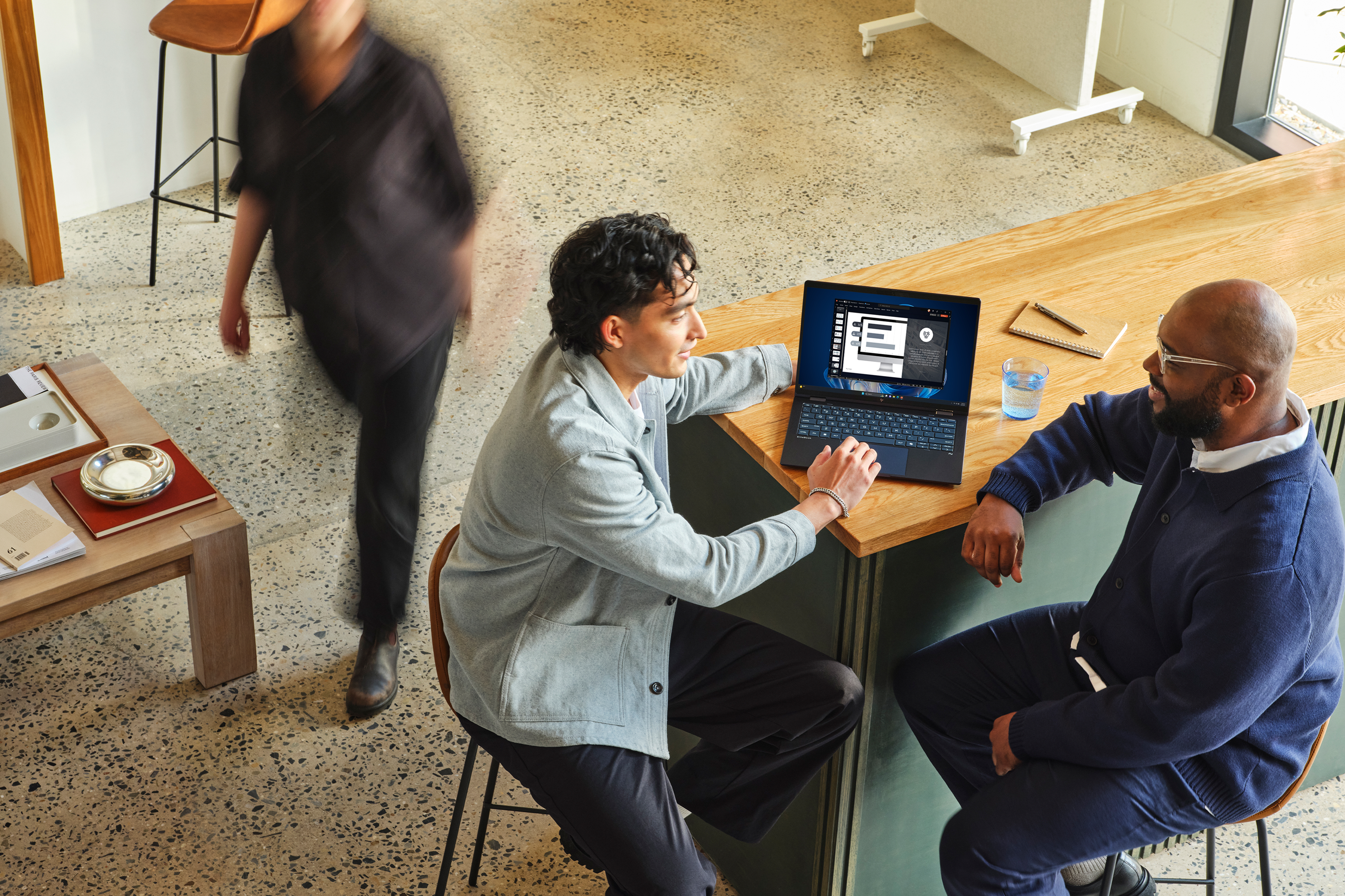 Two office workers sitting at a bar in a meeting area with an open Windows 11 Pro device discussing on-screen content with other furniture in the background