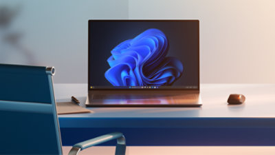 Um dispositivo Windows 11 Pro aberto exibindo uma imagem blue bloom, posicionado sobre uma mesa com mouse, cadeira de escritório e bloco de notas