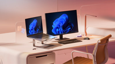 Uma estação de trabalho Windows 11 Pro, um notebook aberto e um monitor grande estendido, ambos exibindo a imagem rainbow bloom, com teclado, mouse, luminária e cadeira de escritório, sobre um fundo em degradê rosa