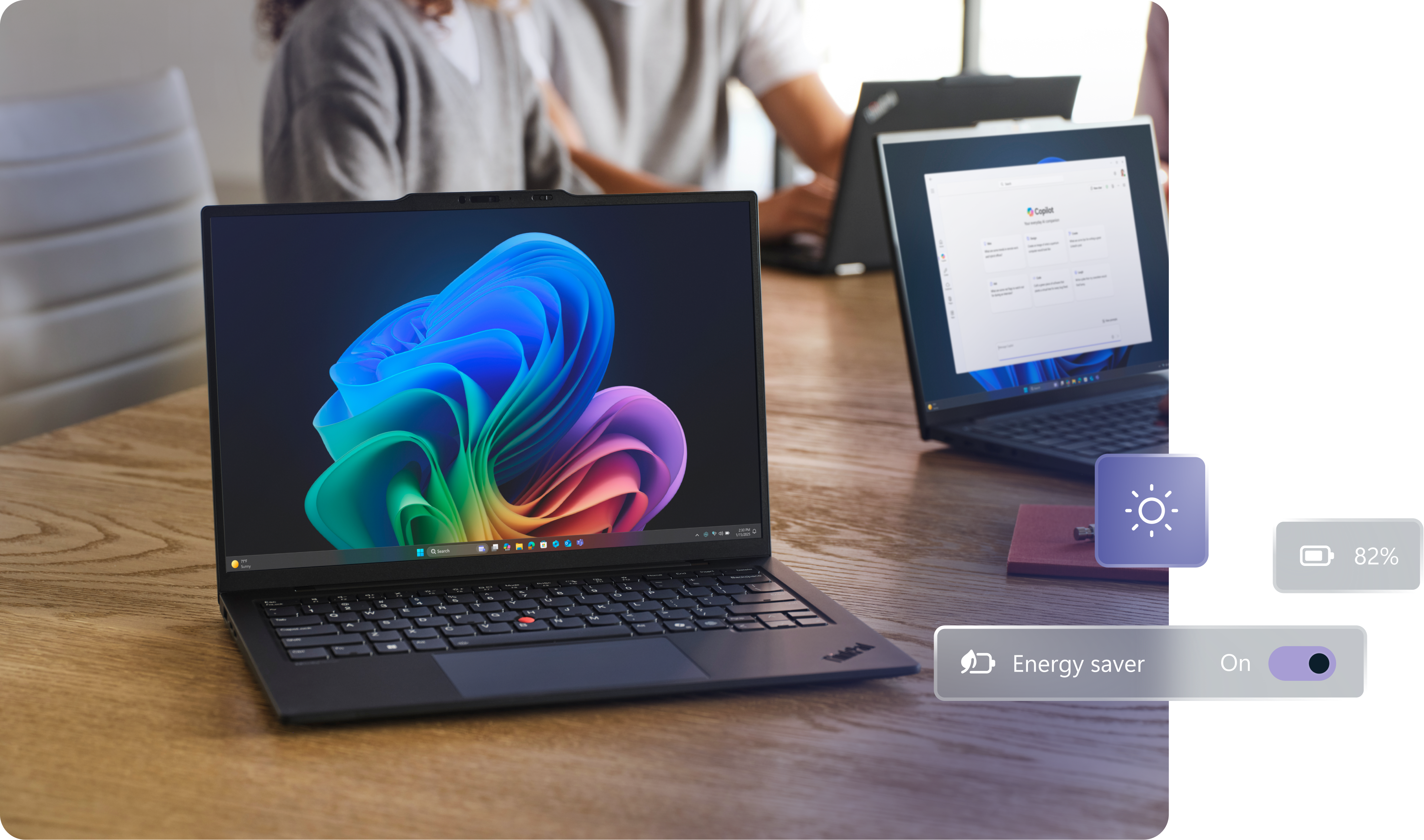 A large worktable with three open Windows 11 Pro devices, one facing forward displaying the rainbow bloom image, with two people working in the background, and conceptual images showing energy use and battery level