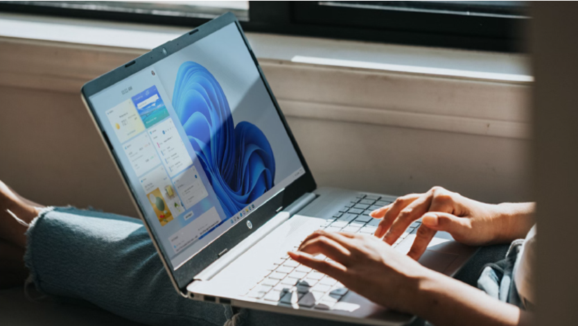 Image of person sitting by a windowsill with a laptop showing the Windows 11 home screen with widgets