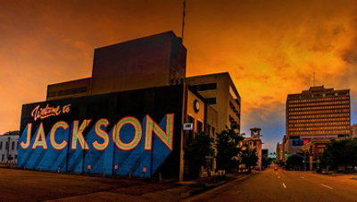 View of a downtown street with a vibrantly colored mural on the closest building reading “Welcome to Jackson”.