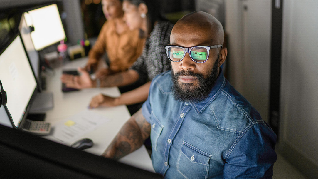 Man in the foreground wearing glasses and a denim shirt looking at a monitor with the glare reflected on his lenses, flanked by two women in the blurred background looking at monitors