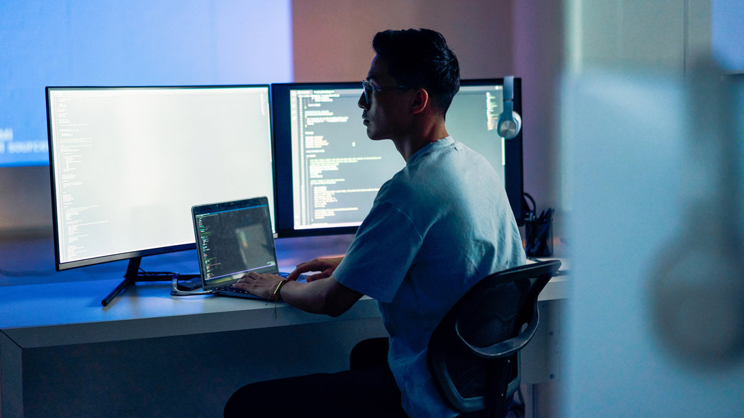 Man sitting in a darkened office wearing glasses as he writes code on a laptop and checks it on dual monitors