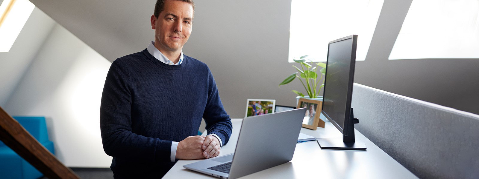 Ein Mann steht an einem Schreibtisch, auf dem ein geöffnetes Windows Laptop-Gerät steht, an dessen Seite sich ein Monitor befindet
