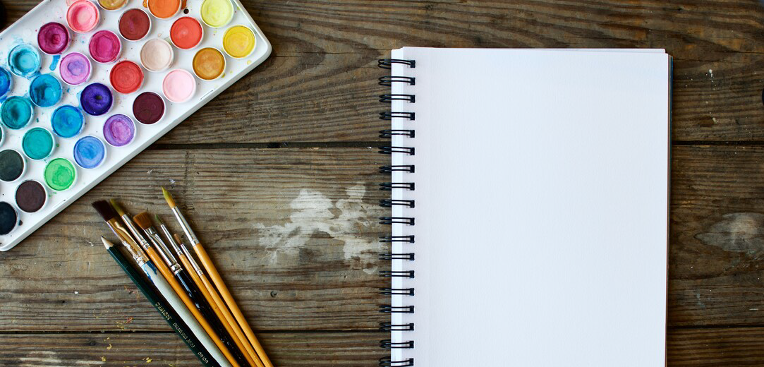 Paint brushes next to an open sketch book and watercolor palette on a wood surface