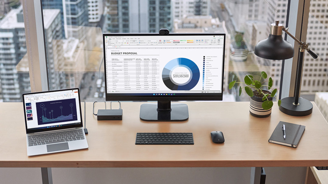 Zařízení Surface zobrazené připojené k Surface Dock 2 na kancelářském stole s externím monitorem, klávesnicí, myší, perem a notebookem poblíž